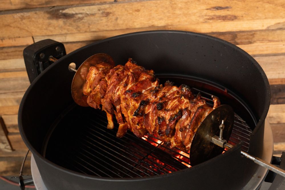 Stacked marinated pork al pastor roasting on a rotisserie skewer over indirect charcoal heat inside a grill.