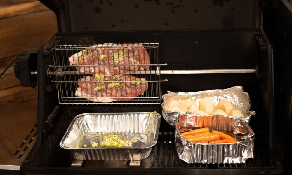 Meat roasting in a rotisserie basket inside a BBQ alongside foil trays of potatoes, carrots and herb butter.