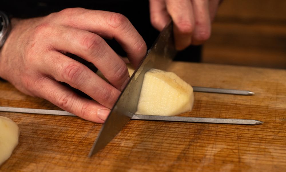 Hands slicing a peeled potato on a wooden board with metal skewers used as cutting guides.