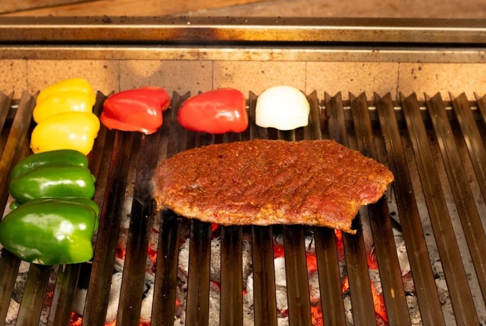 Seasoned steak and halved capsicums (green, yellow, red) along with a white onion grilling over hot coals on a Flaming Coals Parrilla BBQ.