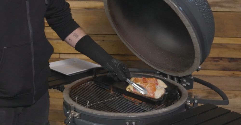 Person using tongs to sear stuffed chicken breasts in a cast iron pan inside a Weber-style kamado grill.